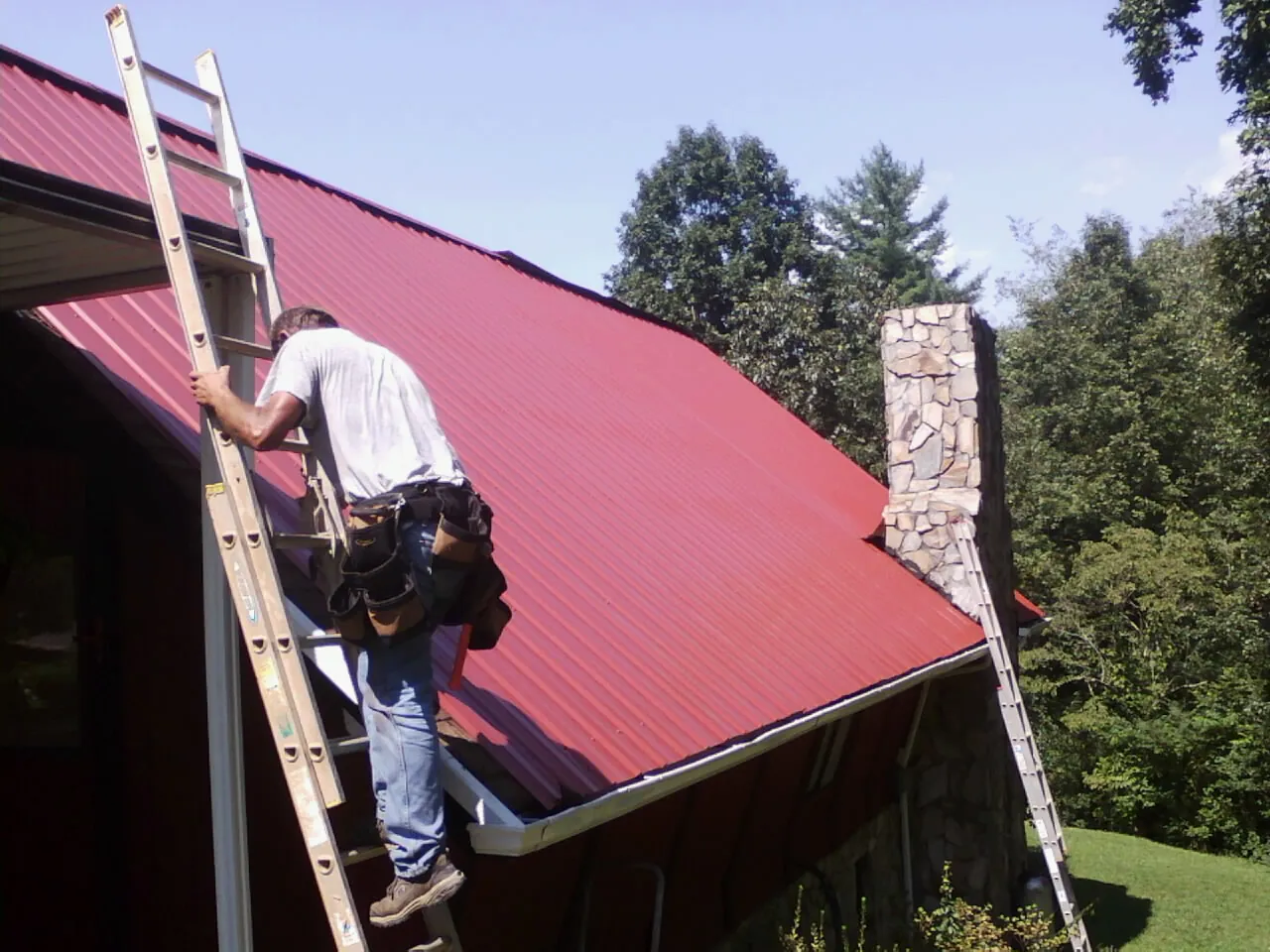 Roofer on a ladder beside a red metal roof and stone chimney — Soffit & Fascia Repair by Roofing Brothers in Prudence Island