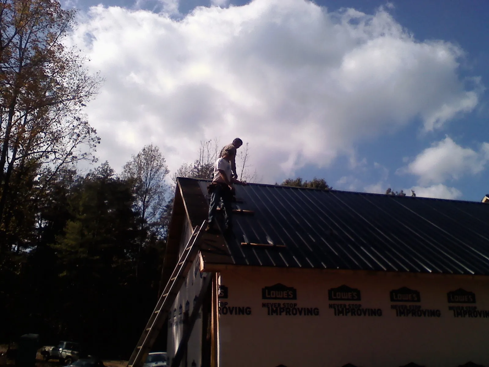 Roofer fastening dark metal panels high against a cloudy sky, from a Roof Inspection job in Prudence Island